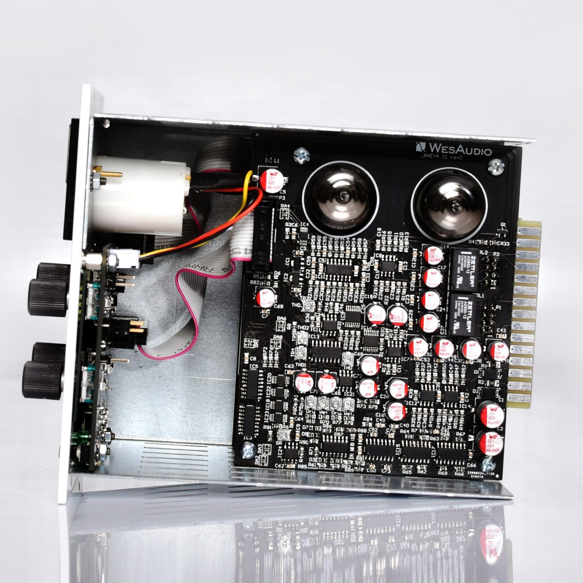 An open electronics component board with visible circuitry, capacitors, connectors, and a motor-like part, photographed on a clean white studio background with a reflective surface below.