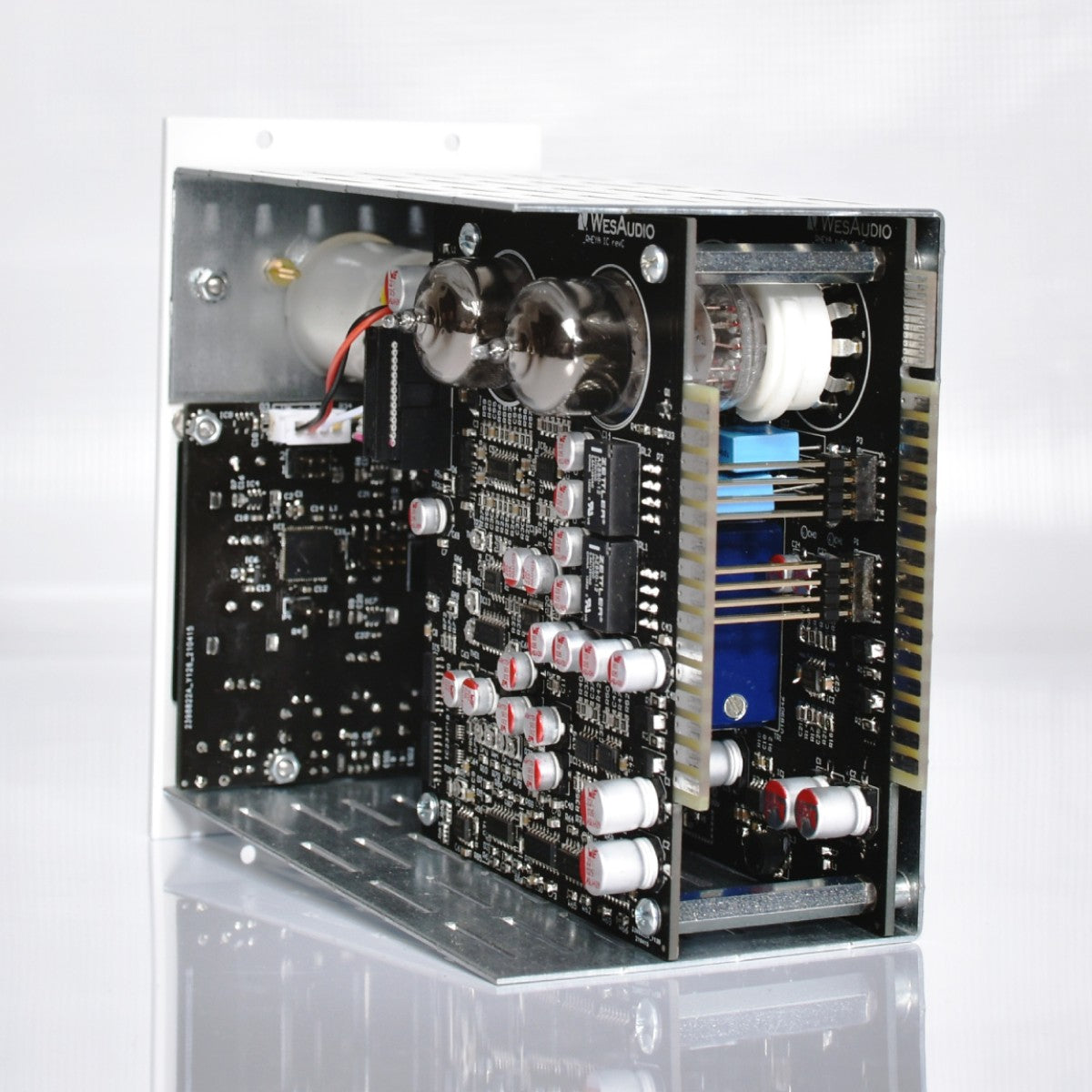 An open metal electronic amplifier or audio equipment chassis showing internal circuit boards, capacitors, and vacuum tubes on a reflective white surface.